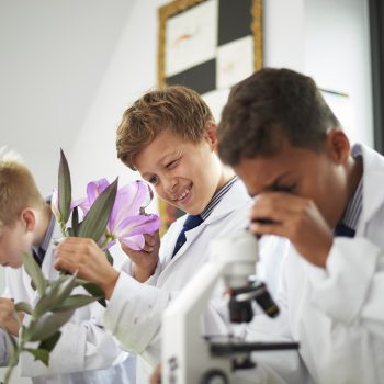 Three school children in science lab coats, looking at flowers and a microscope